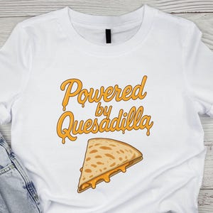 Puede incluir: Camiseta blanca con el texto "Powered by Quesadilla" en escritura naranja sobre una quesadilla de dibujos animados. La quesadilla tiene forma de triángulo con queso derretido.