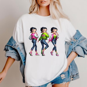 May include: White t-shirt featuring a cartoon illustration of three Betty Boop characters in different outfits. Each character wears a jacket, jeans, and sneakers. The jackets are pink, green, and pink. The jeans are blue, and the sneakers are pink and green.