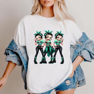 May include: White t-shirt featuring a cartoon design of three Betty Boop-like figures. Each figure wears a green top with "AC/DC" text, black pants, and boots. They also have green headbands and are making a hand gesture.