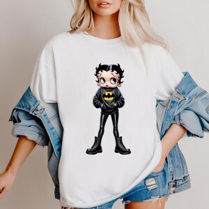 May include: White t-shirt featuring a cartoon Betty Boop character dressed as Batman. The character wears a black sweater with a yellow bat symbol, black pants, and black boots. The image is a pop culture mashup.