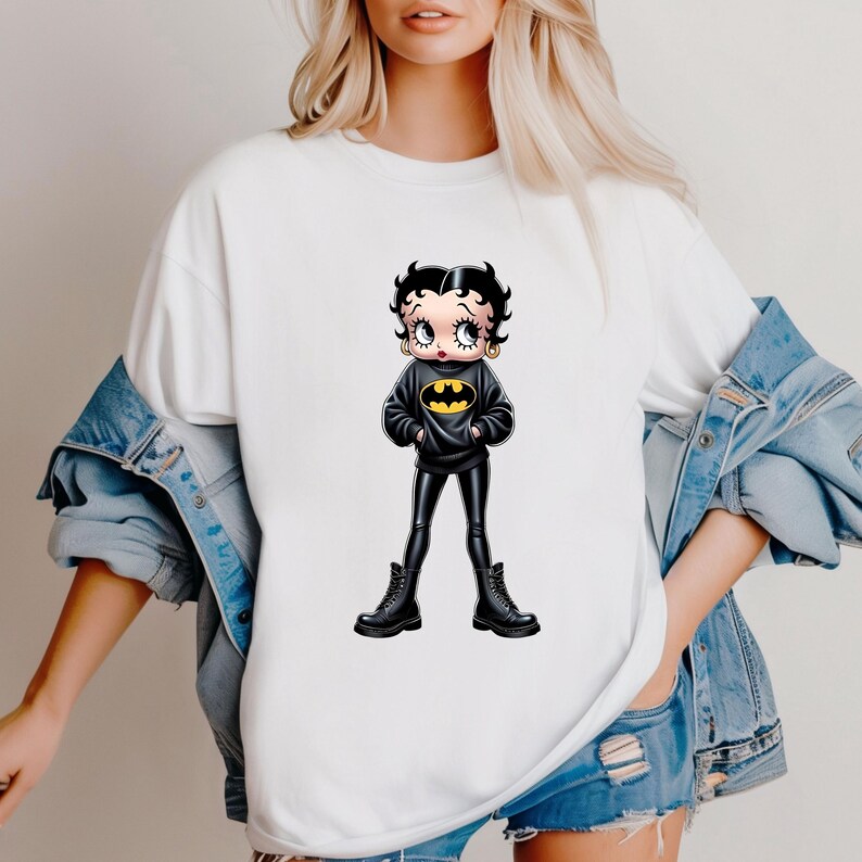 May include: White t-shirt featuring a cartoon Betty Boop character dressed as Batman. The character wears a black sweater with a yellow bat symbol, black pants, and black boots. The image is a pop culture mashup.