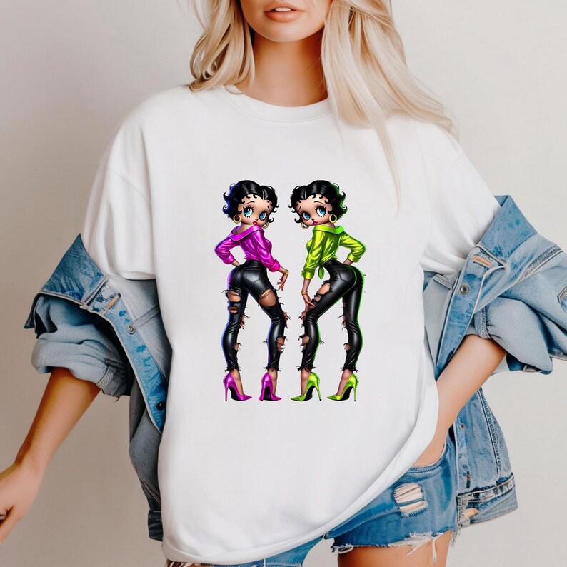 May include: White t-shirt featuring a vibrant graphic of two cartoon women in stylish outfits. The women are depicted in a dynamic pose, wearing ripped black pants, high heels, and colorful tops. The artwork has a bold, eye-catching aesthetic.