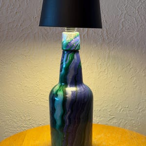 May include: A handcrafted lamp made from a repurposed bottle. The bottle is painted with swirling shades of green, blue, and purple. It is topped with a black lampshade. The lamp sits on a wooden table.