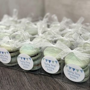 May include: A collection of macarons, each in a clear box tied with white ribbon. The macarons are light green and brown, with a sticker saying "Baby Boy Arriving Soon". The boxes are presented on a wooden surface.
