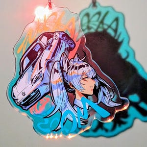 Op de afbeelding: Een heldere, iriserende acryl bedel met een gestileerd anime meisje met blauw haar en een autografiek. De bedel heeft een turquoise omtrek met oranje en blauwe accenten. Het woord "OLA" is zichtbaar in turquoise.