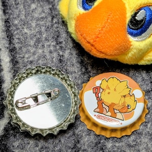 May include: A Chocobo-themed bottle cap badge and a Chocobo plush toy. The bottle cap badge has a silver safety pin on the back and a Chocobo graphic on the front. The plush toy is yellow with blue eyes and an orange beak.