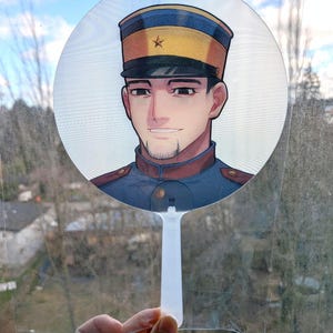 May include: A round, clear plastic fan featuring an anime-style illustration of a man in a military uniform. The man has dark hair and a goatee, and is wearing a hat with a gold star. The fan has a white handle.