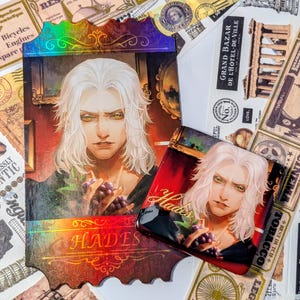 May include: An illustration of a person with white hair and yellow eyes holding grapes. The image is on a decorative card with the word "HADES" and a holographic border. A smaller, similar image is also present.
