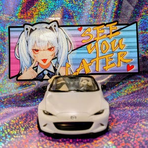 Może przedstawiać: Biały samochodzik Mazda Miata z kierowcą pandą zaparkowany przed naklejką. Naklejka przedstawia dziewczynę w stylu anime z białymi włosami i napisem "See You Later" złotymi literami.