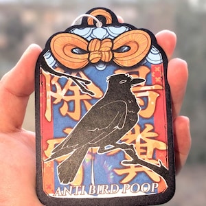 Puede incluir: Una etiqueta rectangular con una ilustración de un cuervo negro posado en una rama. La etiqueta tiene un fondo azul y rojo con caracteres japoneses dorados y naranjas y el texto "ANTI BIRD POOP". Un lazo naranja decora la parte superior.