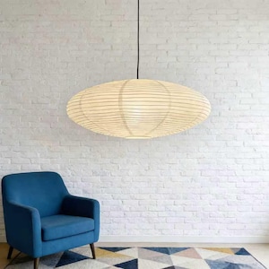 以下が含まれることがあります： クリーム色の紙のようなシェードと細い黒い線が入った、楕円形のペンダントライト。黒いコードから吊り下げられています。青いアームチェアと柄物のラグが下にあり、白いレンガの壁を背景にしています。