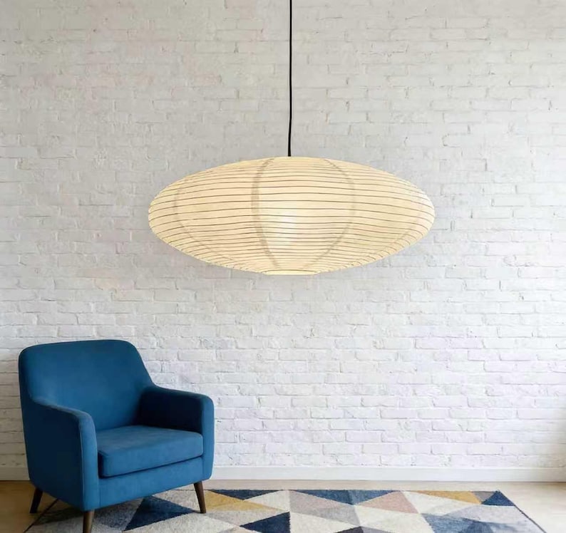 以下が含まれることがあります： クリーム色の紙のようなシェードと細い黒い線が入った、楕円形のペンダントライト。黒いコードから吊り下げられています。青いアームチェアと柄物のラグが下にあり、白いレンガの壁を背景にしています。