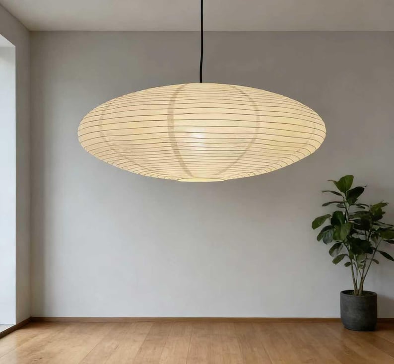 以下が含まれることがあります： クリーム色の紙シェードと黒い水平線が入った楕円形のペンダントライト。黒いコードで吊り下げられており、薄い灰色の壁と木の床がある部屋にあります。鉢植えの植物が隅にあります。スタイリッシュなインテリアデザイン要素です。