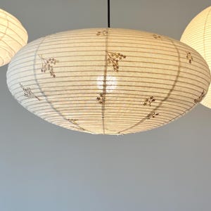 May include: An oval-shaped paper pendant light fixture with a ribbed texture and a delicate brown floral pattern printed across the surface. The lamp is suspended by a thin black cord and emits a soft, warm glow from an internal light source.