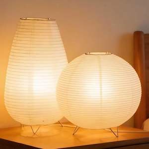 May include: Two illuminated paper lanterns, one teardrop-shaped and the other spherical, sit on a wooden surface. The lamps emit a warm, soft glow, and are supported by delicate metal wire frames. The background includes a wooden headboard.