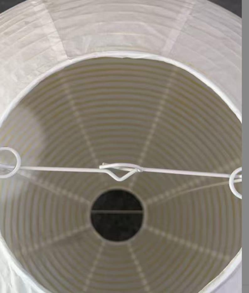 以下が含まれることがあります： 円形の白い紙提灯。白い金属フレームと中央に暗い開口部があります。プリーツ加工された紙がストライプ模様を作り出しています。屋内または屋外での使用に適した装飾品です。