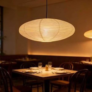 以下が含まれることがあります： 天井から吊り下げられた、大きな楕円形の紙製ランタンが、レストランに温かい光を投げかけています。 ランタンはクリーム色で水平線があり、ダイニングエリアにはお皿とグラスが置かれたテーブルがあります。