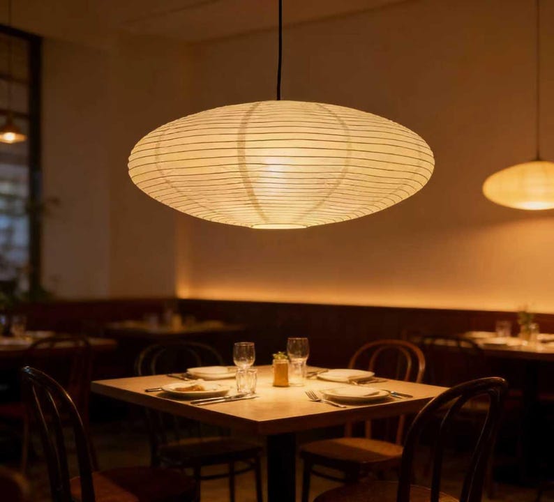 以下が含まれることがあります： 天井から吊り下げられた、大きな楕円形の紙製ランタンが、レストランに温かい光を投げかけています。 ランタンはクリーム色で水平線があり、ダイニングエリアにはお皿とグラスが置かれたテーブルがあります。