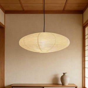 May include: An oval-shaped pendant light fixture with a paper shade hangs from the ceiling. The shade is a warm, off-white color with thin, horizontal lines. The light fixture is suspended in a room with a neutral color palette and wooden accents.