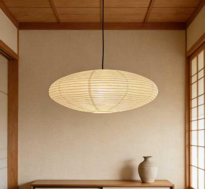 以下が含まれることがあります： 天井から吊り下げられた、楕円形の紙製ペンダントライト。シェードは温かみのあるオフホワイトで、細い水平線が入っています。照明器具は、ニュートラルな色使いと木製のアクセントがある部屋に吊り下げられています。