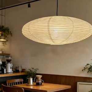 以下が含まれることがあります： 天井から吊り下げられた、大きな楕円形の紙製ランタン。クリーム色で、細い黒い水平線が入っています。カフェのインテリアは柔らかく照らされており、下には木製のテーブルと椅子が見えます。