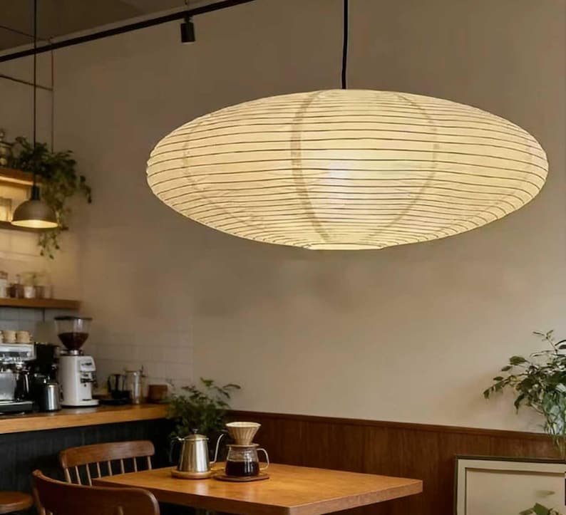 以下が含まれることがあります： 天井から吊り下げられた、大きな楕円形の紙製ランタン。クリーム色で、細い黒い水平線が入っています。カフェのインテリアは柔らかく照らされており、下には木製のテーブルと椅子が見えます。