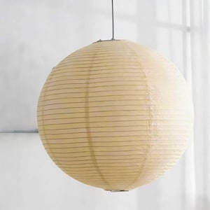 May include: A large, round paper lantern with a cream-colored hue. The lantern is suspended from a thin, dark wire and features horizontal, parallel lines across its surface. The overall design is simple and elegant, suitable for interior lighting.