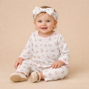Puede incluir: Un beb&eacute; con un conjunto blanco con estampado floral, que incluye una camiseta de manga larga, pantalones y una diadema con un lazo. El conjunto tiene ribetes rosas y se combina con zapatos beige. El beb&eacute; est&aacute; sonriendo.