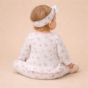 Puede incluir: Un beb&eacute; con un conjunto de pijama blanco con estampado floral con ribete rosa y una diadema a juego. La parte superior y los pantalones de manga larga est&aacute;n cubiertos de peque&ntilde;as flores rosas. La diadema tiene un lazo.