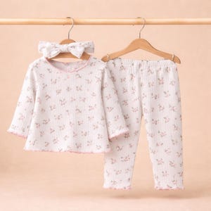 Puede incluir: Un conjunto de pijama blanco con estampado floral, que incluye una camiseta de manga larga, pantalones y una diadema a juego. El conjunto tiene ribetes rosas y est&aacute; colgado de una percha de madera.