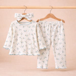 Puede incluir: Un conjunto de pijama color crema con un estampado floral y una diadema a juego. La parte superior de manga larga y los pantalones tienen un ribete azul y cuelgan de una percha de madera. El conjunto est&aacute; hecho de tela acanalada.