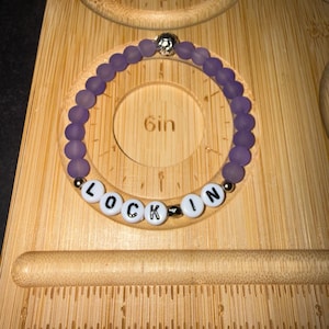May include: A circular beaded bracelet featuring matte purple beads and white letter beads spelling LOCK-IN, accented with small silver-toned metallic beads. The bracelet is positioned on a wooden board with a 6-inch measurement marking.