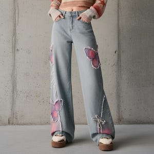 Könnte beinhalten: Eine Frau trägt einen gecroppten, gestreiften Pullover in Rosa, Orange und Beige, kombiniert mit hellblauen Jeans. Die Jeans haben große Schmetterlingsapplikationen in Rosa und Lila und einen weiten Beinverlauf. Das Outfit wird durch Plateauschuhe ergänzt. Die Jeanslänge beträgt ca. 100 cm.