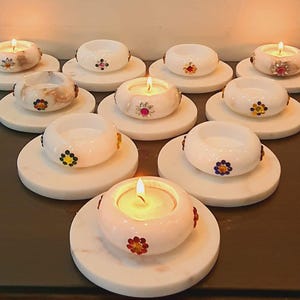 May include: A collection of lit candles in white, decorative holders with colorful floral accents. The candles sit on round, white marble bases, creating a warm and inviting ambiance. The candle holders are approximately 3 inches in diameter.