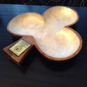 May include: A wooden, clover-shaped dish with a pearlescent interior. The dish has a dark brown border and a small rectangular base with text that reads "Souvenir". The dish is likely a decorative item or a trinket tray.
