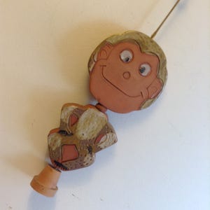 May include: A whimsical ceramic monkey wind chime with a smiling face and tan fur. The monkey's body is a brown and tan pattern. The chime has a small terracotta pot at the bottom.