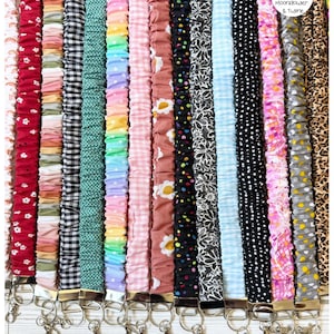 Op de afbeelding: Een collectie van stoffen scrunchie-lanyards in diverse patronen en kleuren, waaronder bloemen, gingham, stippen en luipaardprint. Elke lanyard heeft een geplooid stoffen ontwerp met een zilverkleurige metalen sluiting en ring onderaan.