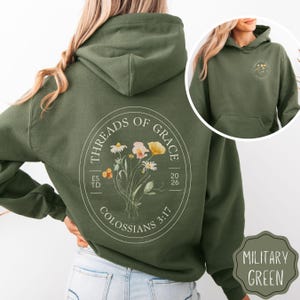 May include: Military green hoodie with a floral design and the text "THREADS OF GRACE" in a circular emblem. The hoodie features a front pocket and a hood. The design includes the text "COLOSSIANS 3:17".
