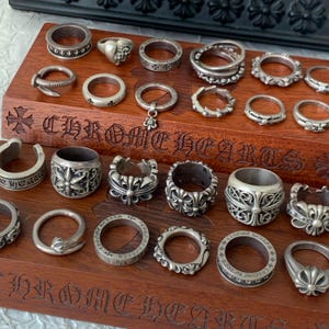 Puede incluir: Una colección de anillos de plata exhibidos en un soporte de madera con el texto Chrome Hearts grabado en el frente. Los anillos presentan varios diseños, incluyendo motivos florales, cruces, bandas lisas y bordes texturizados.