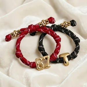 Pode incluir: Duas pulseiras de corda. Uma é vermelha com detalhes brancos, a outra é preta com detalhes brancos. Ambas têm detalhes e berloques em metal dourado. Uma pulseira tem o número "67" e a outra a letra "T".