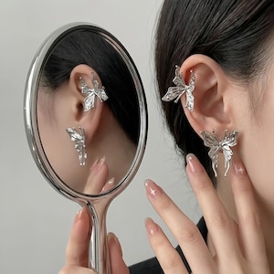 May include: Silver butterfly ear cuffs and earrings. The image shows a person wearing a pair of butterfly-shaped ear cuffs and earrings. The earrings are reflected in a handheld mirror. The jewelry has a detailed, open-wing design.