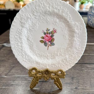 Puede incluir: Un plato decorativo blanco con un patrón floral y de hojas en relieve alrededor del borde. El centro del plato presenta una rosa rosa con hojas verdes. El plato se exhibe en un soporte de metal dorado.