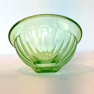 May include: A light green glass bowl with a rounded rim and a geometric, ribbed design. The bowl is made of transparent glass and sits on a white surface. The bowl's design features vertical lines that converge towards the base.