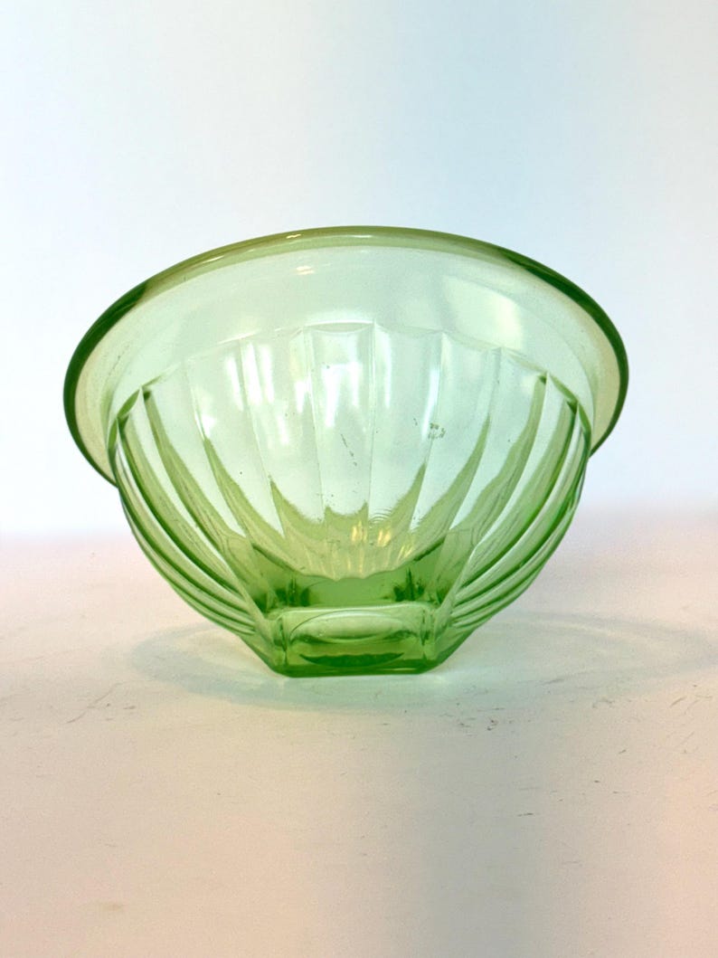 May include: A light green glass bowl with a rounded rim and a geometric, ribbed design. The bowl is made of transparent glass and sits on a white surface. The bowl's design features vertical lines that converge towards the base.