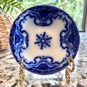 May include: A small, antique white and blue ceramic bowl with a floral design. The bowl is supported by a gold-colored metal stand. The bowl's rim and interior feature intricate blue patterns.
