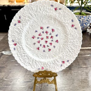 以下が含まれることがあります： 詳細な浮き彫り模様とスカラップエッジが特徴の白い磁器プレート。プレートは、小さなピンクと青の花のクラスターで装飾されています。装飾的な金色のスタンドに展示されており、そのエレガントなデザインを際立たせています。