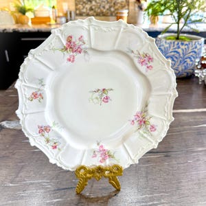 May include: A white, scalloped-edge plate with pink floral designs, displayed on a gold-colored stand. The plate features delicate pink flowers and green leaves, with a central floral motif. The plate is a decorative item.