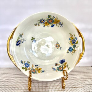 May include: A white porcelain serving dish with gold handles and floral patterns in blue, yellow, and teal. The dish is displayed on a gold-colored stand, showcasing its elegant design.