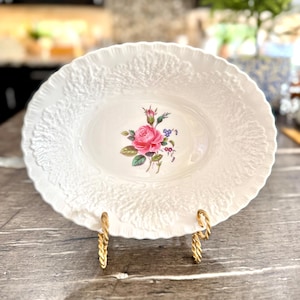May include: White oval serving dish with a floral design featuring a pink rose and blue flowers. The dish has a textured, embossed rim and is displayed on a gold-colored metal stand.