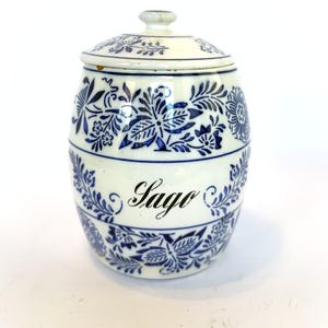 May include: A white ceramic jar with a lid, adorned with blue floral patterns. The jar has the word "Sago" written in elegant script. The design is repeated around the jar's body and lid, creating a cohesive look.